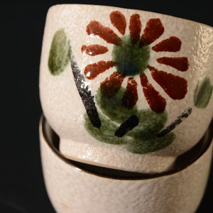 Puede incluir: Dos cuencos de cer&aacute;mica blanca con un dise&ntilde;o floral. El cuenco superior tiene una gran flor roja con hojas verdes y detalles negros. El cuenco inferior es blanco liso.