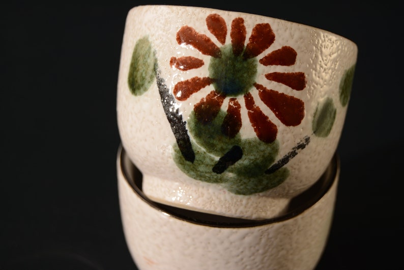 Puede incluir: Dos cuencos de cer&aacute;mica blanca con un dise&ntilde;o floral. El cuenco superior tiene una gran flor roja con hojas verdes y detalles negros. El cuenco inferior es blanco liso.