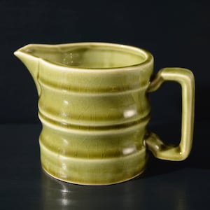 May include: A green ceramic creamer with a textured surface and a rounded handle. The creamer has a ribbed design.
