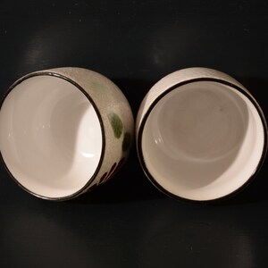 Puede incluir: Dos tazas de cer&aacute;mica blanca con bordes negros y dise&ntilde;os florales verdes y marrones. Las tazas est&aacute;n vac&iacute;as y est&aacute;n sobre una superficie negra.