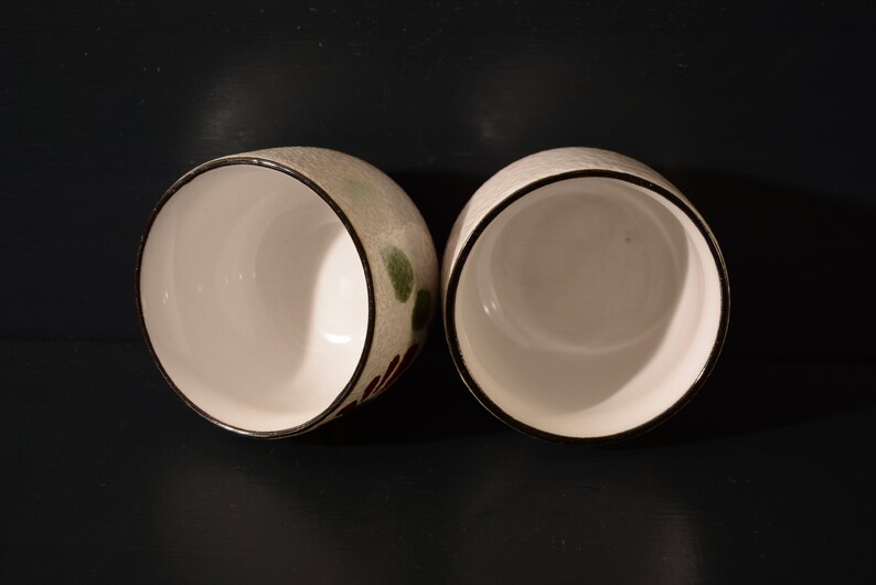 Puede incluir: Dos tazas de cer&aacute;mica blanca con bordes negros y dise&ntilde;os florales verdes y marrones. Las tazas est&aacute;n vac&iacute;as y est&aacute;n sobre una superficie negra.