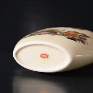 May include: A white ceramic vase with a floral design in shades of pink, red, blue, and green. The vase has a rounded shape and a flat bottom. The bottom of the vase has a red label that reads "Made in Japan".