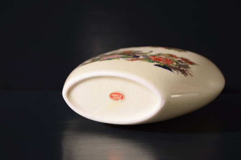 May include: A white ceramic vase with a floral design in shades of pink, red, blue, and green. The vase has a rounded shape and a flat bottom. The bottom of the vase has a red label that reads "Made in Japan".