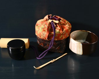 Op de afbeelding: Een traditionele Japanse theeceremonie set met een geweven bamboe theedoos, een zwarte gelakte theedoos, een houten theewisker en een bruin en witte keramische theekom.