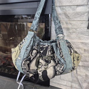 May include: A handmade tote bag with a unique design. The bag features a black and white print with a woman's face, and is accented with blue and olive green patterned fabric. The bag has a matching blue strap and white drawstring ties.