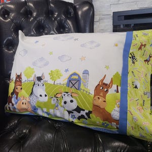 May include: A rectangular pillowcase with a farmyard design, showing cartoon animals like horses, cows, and a donkey. The main design is on a white background with a green and blue border. The pillowcase is on a black leather chair.