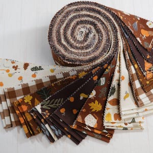 May include: A rolled bundle of fabric strips in autumnal colours. The fabric features patterns of leaves, pumpkins, and gingham checks. The colours include brown, cream, orange, and dark brown. The fabric is likely for quilting or other craft projects.