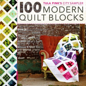 May include: A book cover for "100 Modern Quilt Blocks" by Tula Pink. A vibrant quilt with square designs in multiple colours is draped over a red armchair. The backdrop includes a brick wall.