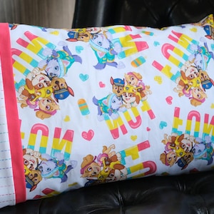 May include: A white pillowcase with a repeating pattern of cartoon puppies, ice lollies, and hearts. The design includes the text "WOW" in pink and yellow. The pillowcase has a pink border and a white section with rainbow stitching.