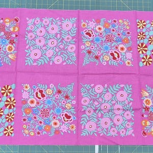 May include: A magenta fabric panel with eight square floral designs. Each square features a unique arrangement of flowers in pink, red, yellow, blue, and green. The fabric is laid out on a green cutting mat.