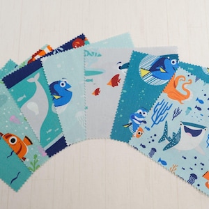 Può includere: Un ventaglio di sette pezzi di tessuto quadrati con vari motivi a tema acquatico. I disegni presentano pesci dei cartoni animati, balene e altre creature marine in tonalità di blu, arancione e bianco. Il tessuto ha uno sfondo azzurro.