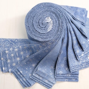 May include: A jelly roll of fabric strips in shades of dusty blue, featuring a variety of silver metallic patterns including swirls, stars, and dots. The fabric is rolled into a tight cylinder with several strips fanned out to display.