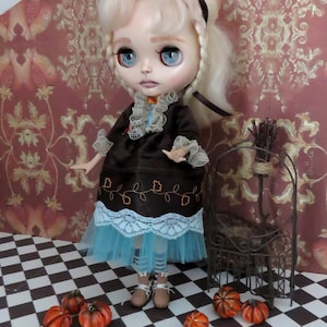 Puede incluir: Una muñeca con cabello rubio y ojos azules, vestida con un vestido marrón con una falda azul y ribete de encaje blanco. Pequeñas calabazas naranjas están esparcidas en un suelo a cuadros blanco y negro. Una jaula decorativa de metal está a la derecha.