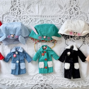 May include: A collection of handmade doll clothes and accessories, including coats in red, blue, and black, and bunny-eared hats in various colors. The coats feature faux fur pockets and floral accents, while the hats have embroidered faces.