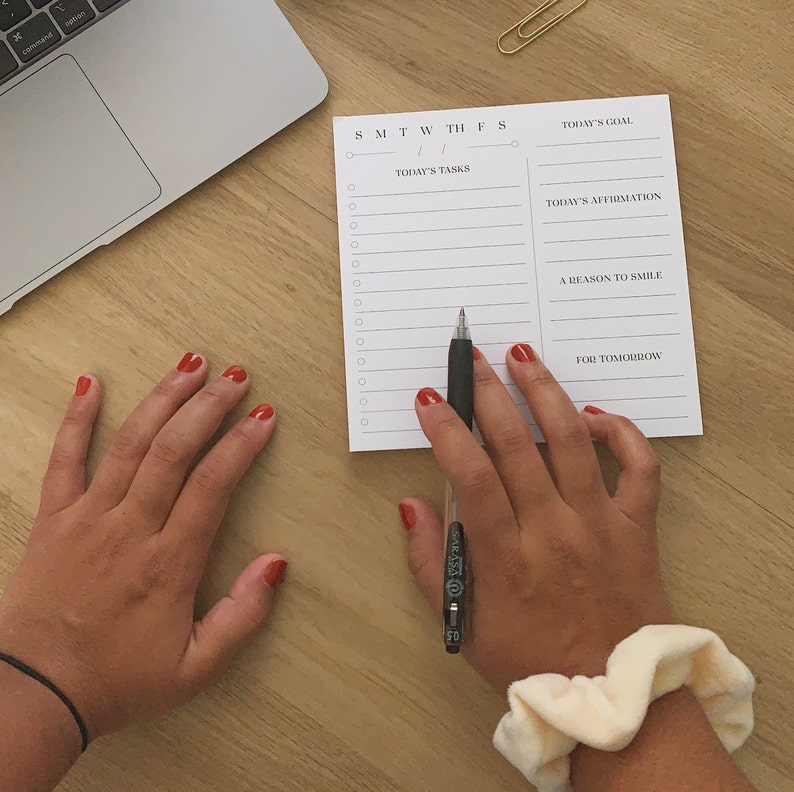 May include: A white notepad with a daily planner layout. The notepad has sections for today's goal, today's tasks, today's affirmation, a reason to smile, and for tomorrow. The notepad is on a wooden surface with a black pen and a hand holding the pen.