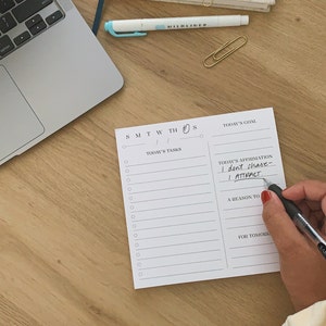 May include: A white daily planner with a black grid and the days of the week labeled. The planner has sections for today's tasks, today's goal, today's affirmation, a reason to, and for tomorrow. The planner is on a wooden surface.