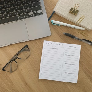 May include: A white daily planner with a checklist for tasks, goals, affirmations, and a reason to smile. The planner has a simple design with black text and a black border.