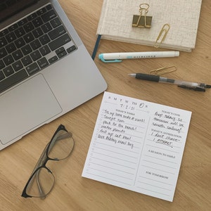 May include: A white notepad with a daily planner and to-do list. The notepad has a black and white design with the days of the week listed at the top. The notepad is on a wooden surface with a laptop, glasses, and a pen.