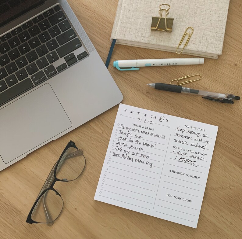 May include: A white notepad with a daily planner and to-do list. The notepad has a black and white design with the days of the week listed at the top. The notepad is on a wooden surface with a laptop, glasses, and a pen.