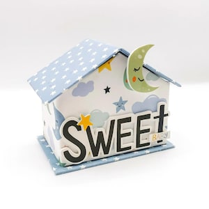 May include: A blue and white house-shaped box with a moon on the roof. The box has a white background with blue clouds and stars. The word "SWEET" is written in black letters on the front of the box, with the words "BABY" in smaller letters below.