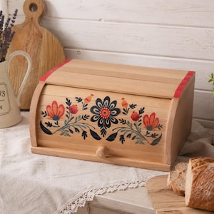 Könnte beinhalten: Ein Brotkasten aus hellem Holz mit einem Rolldeckel, der mit einem handgemalten Blumenmuster in Rot, Blau und Grün verziert ist. Die Vorderseite des Kastens hat einen kleinen Holzknauf zum Öffnen und die Oberkanten sind mit roten Linien akzentuiert.