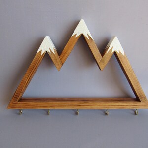 Puede incluir: Estante de pared de madera con forma de monta&ntilde;a con acabado de madera natural y picos pintados de blanco. El estante tiene una fila de peque&ntilde;os ganchos en la parte inferior. Una pieza decorativa y funcional para organizar llaves o objetos peque&ntilde;os.