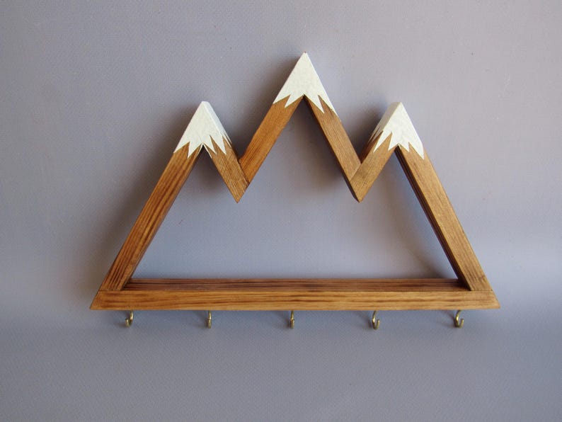 Puede incluir: Estante de pared de madera con forma de monta&ntilde;a con acabado de madera natural y picos pintados de blanco. El estante tiene una fila de peque&ntilde;os ganchos en la parte inferior. Una pieza decorativa y funcional para organizar llaves o objetos peque&ntilde;os.