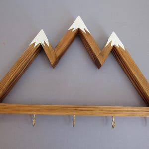 Puede incluir: Estante de pared de madera con forma de monta&ntilde;a con tres picos, cada uno pintado de blanco. La parte inferior tiene cuatro ganchos de lat&oacute;n. La madera tiene un tono c&aacute;lido y natural. Ideal para colgar objetos.