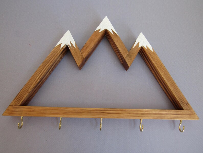 Puede incluir: Estante de pared de madera con forma de monta&ntilde;a con tres picos, cada uno pintado de blanco. La parte inferior tiene cuatro ganchos de lat&oacute;n. La madera tiene un tono c&aacute;lido y natural. Ideal para colgar objetos.