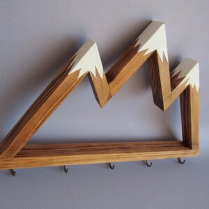 Puede incluir: Estante de pared de madera con forma de monta&ntilde;a, con acabado de madera natural y picos pintados de blanco. El estante tiene una base rectangular con cinco peque&ntilde;os ganchos para colgar objetos.