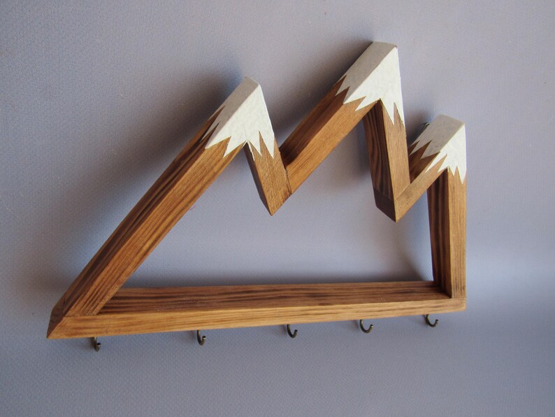 Puede incluir: Estante de pared de madera con forma de monta&ntilde;a, con acabado de madera natural y picos pintados de blanco. El estante tiene una base rectangular con cinco peque&ntilde;os ganchos para colgar objetos.