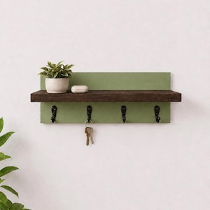 Könnte beinhalten: Ein wandmontierter Flur-Organizer mit einem dunkelbraunen Holzregal auf einer rechteckigen, salbeigrünen Rückwand. Das Element verfügt über vier schwarze Metallhaken zum Aufhängen von Gegenständen, wobei ein Schlüsselbund an einem Haken hängt.