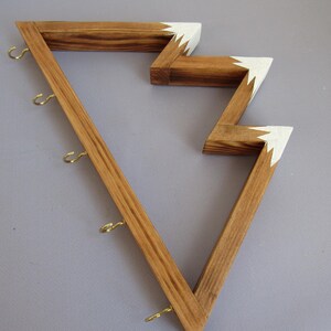 Puede incluir: Estante de pared de madera con un dise&ntilde;o en zigzag &uacute;nico. El estante est&aacute; hecho de madera marr&oacute;n claro con detalles blancos en los bordes. Cuenta con varios ganchos peque&ntilde;os de color dorado para colgar objetos.