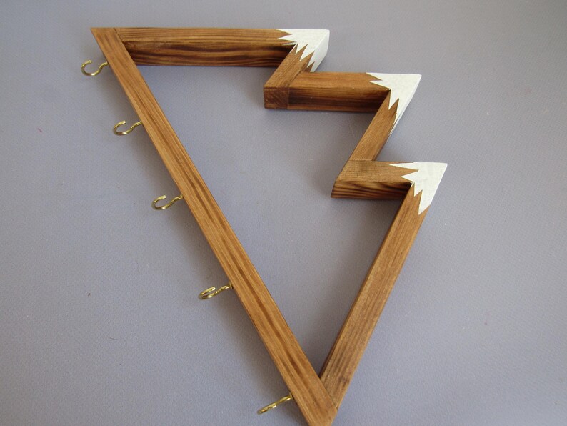Puede incluir: Estante de pared de madera con un dise&ntilde;o en zigzag &uacute;nico. El estante est&aacute; hecho de madera marr&oacute;n claro con detalles blancos en los bordes. Cuenta con varios ganchos peque&ntilde;os de color dorado para colgar objetos.
