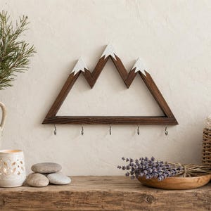 Puede incluir: Estante de pared de madera con forma de monta&ntilde;a, con marco marr&oacute;n oscuro y picos pintados de blanco. El estante tiene cinco ganchos de metal debajo. Se exhibe sobre una superficie de madera r&uacute;stica.