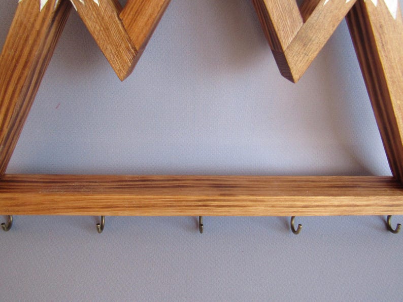 Puede incluir: Llavero de madera con forma de monta&ntilde;a con un estante y seis ganchos de metal. El marco es de madera marr&oacute;n claro con detalles blancos. El llavero est&aacute; montado en una pared gris.