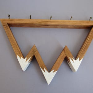 Puede incluir: Estante de pared de madera con dise&ntilde;o de monta&ntilde;a. La madera marr&oacute;n claro tiene detalles blancos pintados en las cimas. Cuenta con cuatro ganchos met&aacute;licos para colgar objetos.