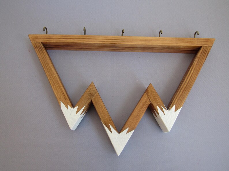 Puede incluir: Estante de pared de madera con dise&ntilde;o de monta&ntilde;a. La madera marr&oacute;n claro tiene detalles blancos pintados en las cimas. Cuenta con cuatro ganchos met&aacute;licos para colgar objetos.