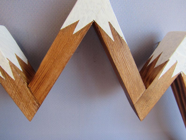 Puede incluir: Adorno de pared de madera con forma de cordillera. Las cimas est&aacute;n pintadas de blanco y la base es de color madera natural. El dise&ntilde;o es en zigzag, creando una silueta de monta&ntilde;a. Esta pieza decorativa es adecuada para un hogar r&uacute;stico o moderno.