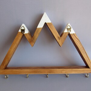Puede incluir: Estante de pared de madera con forma de monta&ntilde;a, con acabado de madera natural y picos pintados de blanco. El estante tiene cinco ganchos de lat&oacute;n a lo largo del borde inferior para colgar objetos. Incluye dos colgadores triangulares de metal en la parte trasera.