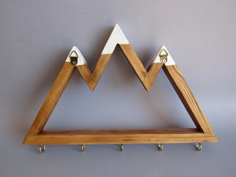 Puede incluir: Estante de pared de madera con forma de monta&ntilde;a, con acabado de madera natural y picos pintados de blanco. El estante tiene cinco ganchos de lat&oacute;n a lo largo del borde inferior para colgar objetos. Incluye dos colgadores triangulares de metal en la parte trasera.
