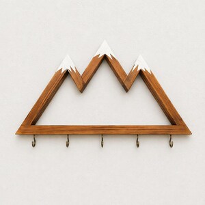 Puede incluir: Perchero de pared de madera con forma de monta&ntilde;a con cinco ganchos de color bronce. Los picos de las monta&ntilde;as est&aacute;n pintados de blanco, contrastando con la madera marr&oacute;n. Un art&iacute;culo decorativo y funcional.