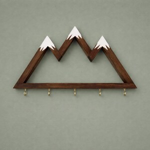 Puede incluir: Estante de pared de madera con forma de monta&ntilde;a con tres picos, pintados de blanco en la parte superior. El estante es de color marr&oacute;n oscuro y tiene seis ganchos de lat&oacute;n en la parte inferior. Est&aacute; montado en una pared verde apagado.