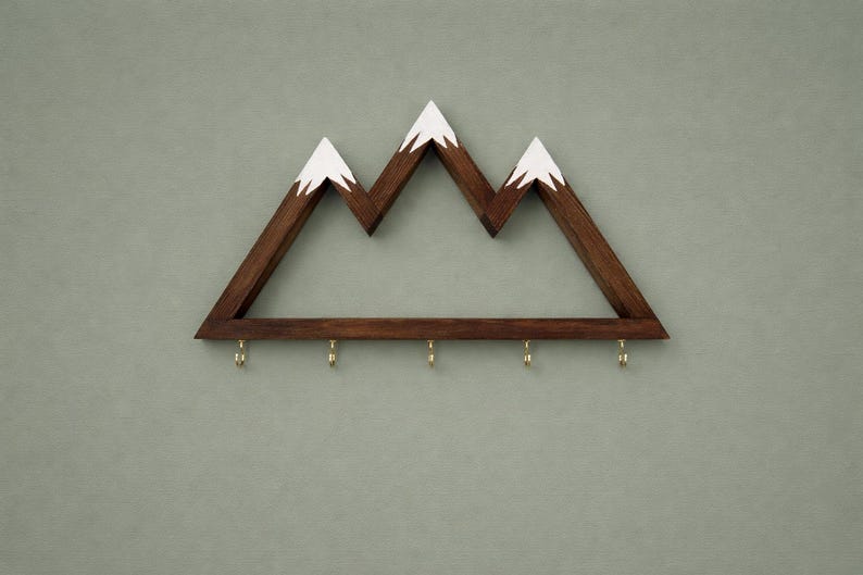 Puede incluir: Estante de pared de madera con forma de monta&ntilde;a con tres picos, pintados de blanco en la parte superior. El estante es de color marr&oacute;n oscuro y tiene seis ganchos de lat&oacute;n en la parte inferior. Est&aacute; montado en una pared verde apagado.