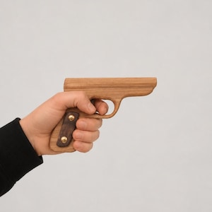 May include: A wooden toy pistol crafted from light-toned wood with a darker wood grip panel secured by two brass-coloured metal rivets. The object features a smooth, rounded barrel and a curved trigger guard, showcasing a simple, solid construction.