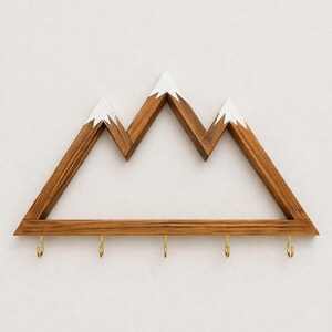 Puede incluir: Estanter&iacute;a de pared con forma de monta&ntilde;a de madera con acabado de madera natural y picos pintados de blanco. La estanter&iacute;a tiene cinco ganchos de color lat&oacute;n en la parte inferior, perfectos para colgar llaves o objetos peque&ntilde;os. Una pieza decorativa y funcional.