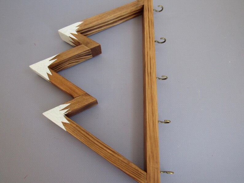 Puede incluir: Llavero de madera con dise&ntilde;o en zigzag y secci&oacute;n vertical con ganchos. La parte en zigzag est&aacute; te&ntilde;ida de marr&oacute;n con detalles blancos en las puntas. La secci&oacute;n vertical es de un tono de madera m&aacute;s claro y tiene varios ganchos met&aacute;licos.