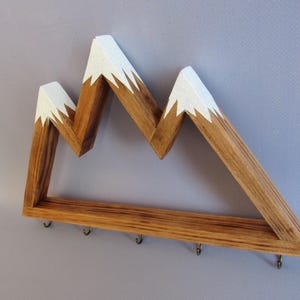 Puede incluir: Estante de pared de madera con forma de monta&ntilde;a, con acabado de madera natural y picos pintados de blanco. El estante tiene una fila de peque&ntilde;os ganchos en la parte inferior. Un art&iacute;culo decorativo y funcional.