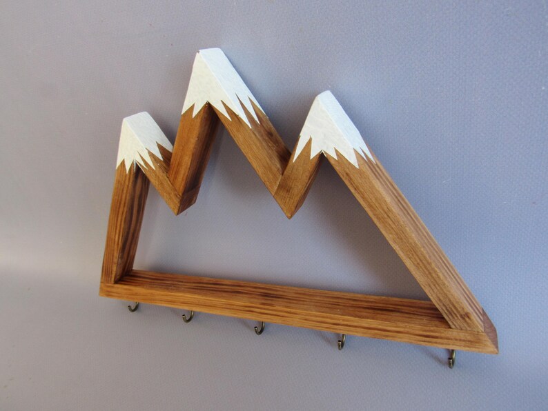 Puede incluir: Estante de pared de madera con forma de monta&ntilde;a, con acabado de madera natural y picos pintados de blanco. El estante tiene una fila de peque&ntilde;os ganchos en la parte inferior. Un art&iacute;culo decorativo y funcional.