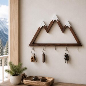 Puede incluir: Un llavero de madera con forma de monta&ntilde;as, con tres picos pintados de blanco. El marco marr&oacute;n oscuro tiene ganchos para llaves, con varios llaveros colgando. Se ve un llavero en forma de casa peque&ntilde;a.